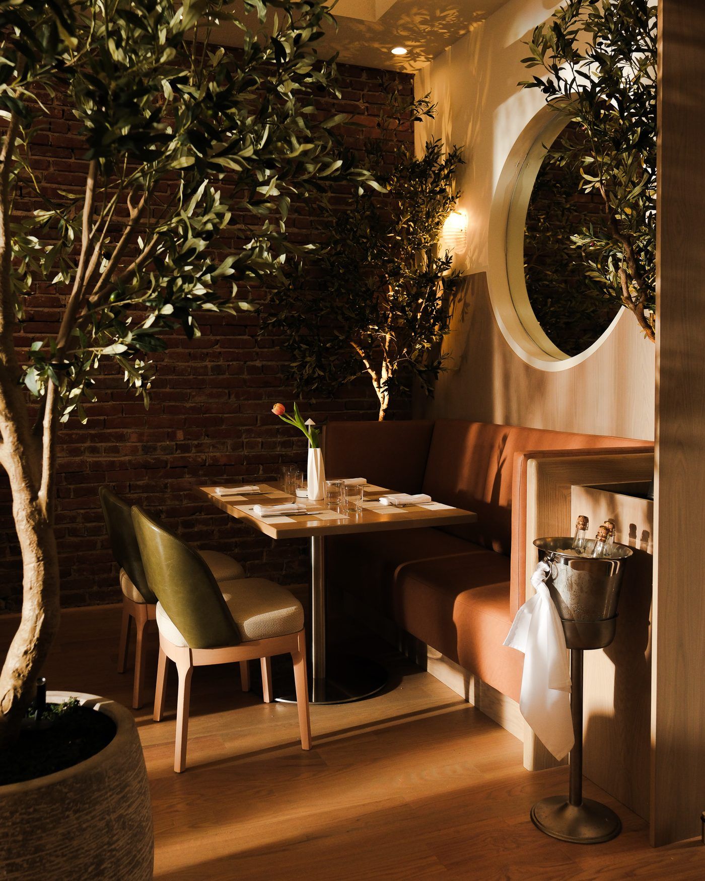 A corner banquette at Clementina beneath an olive tree, with an arched mirror and exposed brick